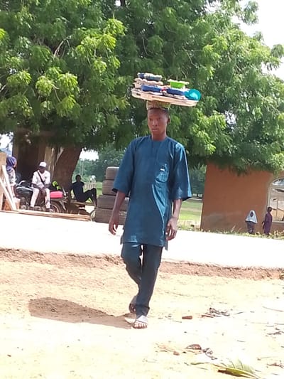A young man walks outdoors, balancing a large tray of items on his head. He is wearing a dark blue tunic and pants, with sandals on his feet. The tray on his head contains various items, including what appear to be packaged goods, some in blue and white wrapping, and a green bowl.

The scene appears to be a casual outdoor setting in a village or rural area. In the background, there is a large tree providing shade, a building with a reddish-brown wall and a window, and several people. To the left of the man, a person sits on a motorcycle next to a structure that looks like it might be a market stall or temporary shelter. Further in the background, another person is seated, and there are stacks of tires. Two small figures, possibly children, are visible near the building on the right.

The lighting suggests it is daytime, likely under sunny conditions given the bright lighting and shadows cast on the ground. The ground is dusty and appears to be unpaved. The overall impression is one of daily life in a West African community. No visible text is present in the image.