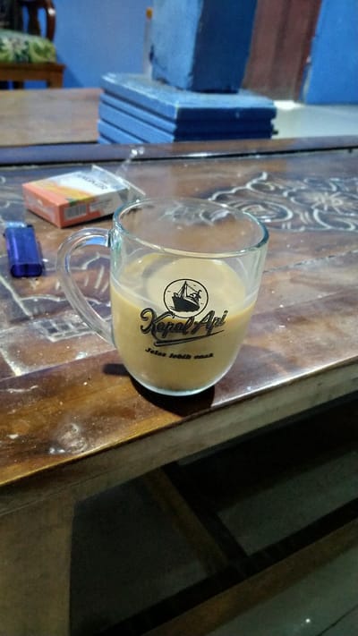 A glass mug filled with a light brown liquid, presumably coffee, sits on a wooden table. The mug has a black logo with a ship and text that reads "Kapal Api" and "Jelas lebih enak". To the left of the mug is a blue lighter and a pack of cigarettes. The table surface has a carved pattern. In the background, a stack of blue objects is visible, as well as part of a chair and a wall. The overall scene suggests a casual setting, likely indoors. The presence of a cigarette pack and lighter implies smoking might be occurring or has recently occurred. There are no people visible in the image. The exact location within Banyusri, Indonesia, cannot be determined from the visual information provided.