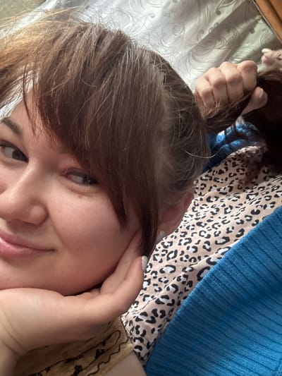 The image is a close-up, selfie-style photograph of a young woman. She has brown hair, styled with bangs that partially cover her forehead and a section of her hair is pulled back by her hand. Her left hand is resting on her chin, supporting her face. She is looking towards the left side of the frame with a slight smile. The background appears to be indoors, with a textured surface that could be bedding or a blanket. There is also a hint of a blue, ribbed garment visible. The image does not provide any specific clues about the location within the city of Маркидоновка, Russia.