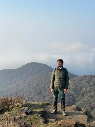 A man is standing on the edge of a mountain in Lwang Ghalel, Nepal. He is wearing a green puffer jacket and green pants. The mountain is covered in trees and bushes. The sky is partly cloudy.