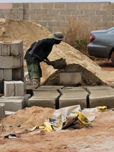 A construction site in Nwofia, Nigeria, presents an adult male engaged in manual labor. The individual, attired in a faded gray cap, dark short-sleeved top, and green trousers, is captured mid-action, operating a dark-colored shovel to transfer dark, granular material from a large mound into a smaller, lower pile on the ground. A thin, light-colored object is visible protruding from his mouth, and a blue band is on his right wrist. To the left of the individual are multiple light-gray concrete blocks stacked vertically. Further right and extending into the mid-ground are numerous rows of similar concrete blocks laid horizontally. On the reddish-brown, sandy foreground are two crumpled bags, one predominantly white with blue/green text, the other yellow. A substantial pile of tan sand occupies the background, partially obscuring the rear section of a gray sedan on the far right. A light-colored concrete block wall with a reddish-brown upper edge is visible in the distant background, accompanied by sparse, dry vegetation.