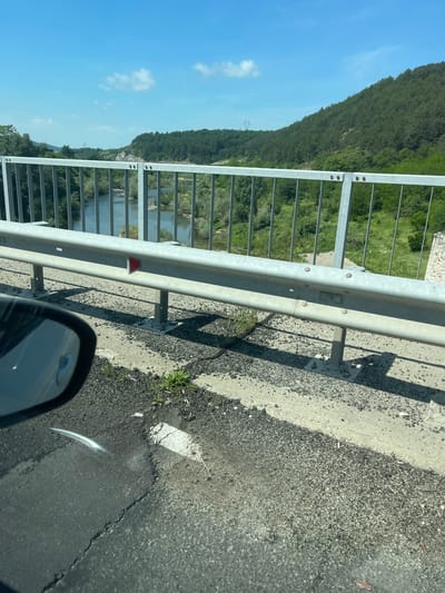 The image, taken from the perspective of a vehicle, captures a scenic view from a road or bridge overlooking a river valley in the area of Balabanovo, Bulgaria. In the bottom right foreground, a portion of a car's side mirror is visible, suggesting an ongoing journey. The immediate right of the frame shows an asphalt road surface meeting a concrete shoulder, both bathed in bright sunlight which casts sharp shadows from the guardrail.

A prominent metal guardrail with vertical bars extends across the mid-ground, separating the road from the vista beyond. A small, red, triangular reflector is attached to the guardrail. Beyond this barrier, a tranquil river winds through a valley, its banks lush with green vegetation and trees.

In the background, a large, verdant hill or mountain, densely covered in trees, rises majestically under a clear blue sky punctuated by a few wispy white clouds. A power line tower is faintly visible on the distant hillside. The bright sunlight and distinct shadows suggest it is daytime, likely late morning or early afternoon, with clear weather conditions. No specific text is visible in the image.