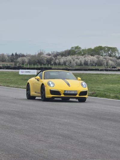 A yellow Porsche sports car with the license plate "CB 0911 XO" is captured on a gray asphalt race track. The track is bordered by a green grassy area and a line of trees with white blossoms. A sign with "TUNING BAZAR" is also visible. The location is identified as the city of Kaloyanovo, Bulgaria. No people are visible in the image.