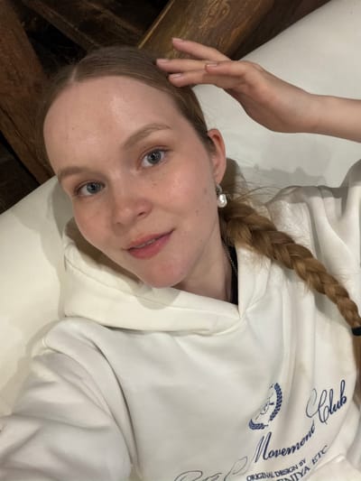 A young person, likely female, with fair skin, light-colored eyes, and light brown hair styled in a braid, is depicted in this indoor portrait. She is lying down, possibly on a bed or couch, against a light-colored surface. A wooden surface is visible in the background, to the left. She is wearing a white hooded sweatshirt with blue text and a small blue emblem on the front. Visible text on the sweatshirt includes words like "Club", what appears to be "Lemon" or "Moment", another cursive word that could be "Moments" or "Mornin'", and the phrase "ORIGINAL DESIGN BY NIYA ETC". The emblem resembles a stylized head within a wreath.
Her right hand is raised and resting near her head. A pearl earring is visible in her right ear, and a thin silver chain is seen around her neck. She looks directly at the camera with a relaxed expression and a slight smile. The image is taken from a slightly overhead perspective. There are no specific visual cues within the image to indicate the time of day or the location of Yus'ki, Russia, other than the provided context.