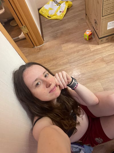A person with long brown hair, wearing a black top and red shorts, is lying on a light-colored surface, possibly a bed or sofa. The person is looking at the camera with their hand near their face.
In the background, there is a wooden floor, a cardboard box with some printed text (including the words "CARTON SIZE" and color options), a yellow plastic bag, and a child's toy cube. The scene appears to be indoors.
There is no indication of the location being in Usol'e, Russia.