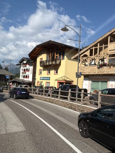The image shows a street scene in Campitello di Fassa, Italy. In the foreground, a black car is driving on the right side of the road. In the middle ground, several parked cars are visible behind a wooden fence. The buildings lining the street are typical of the region, with wooden balconies and decorative facades. One building has a sign that reads "FPB CASSA". In the background, mountains are visible under a partly cloudy sky. The overall impression is of a well-maintained town with a mix of residential and commercial buildings.