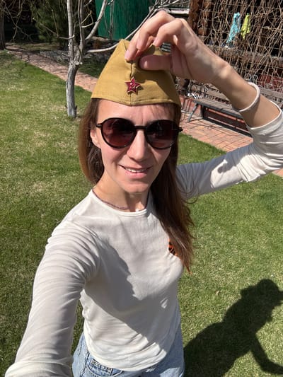 A woman is photographed outdoors on a sunny day. She is wearing sunglasses, a long-sleeved shirt, and jeans. On her head, she wears a military-style cap with a red star. She is smiling and appears to be taking a selfie. The background includes grass, trees, and a brick building with a fence. The photo was taken in the city of Smolenshchina, Russia.