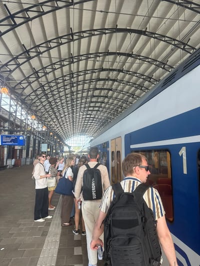 The image depicts a crowded train platform at Haarlem Station in the city of Haarlem, Netherlands. The scene is dominated by the arched, industrial roof structure of the station, which is supported by a network of metal beams. Natural light streams in from the open ends of the platform and through the translucent roofing material.
A blue and white train is visible on the right side of the frame, numbered "1". Several people are standing on the platform, waiting to board or disembark the train. They are diverse in age and gender, dressed in casual summer attire. Many are carrying backpacks and bags.
In the foreground, a man wearing sunglasses and a striped shirt with a black backpack is facing away from the camera, looking towards the train. To his left, another man in a light-colored sweatshirt and black pants is also looking towards the train. A group of people are clustered further down the platform, some in conversation, others looking at their phones. A digital display board showing train information is visible on the left side of the platform. The ground of the platform is paved with brick-like tiles. The overall impression is of a busy transit hub on a bright day.