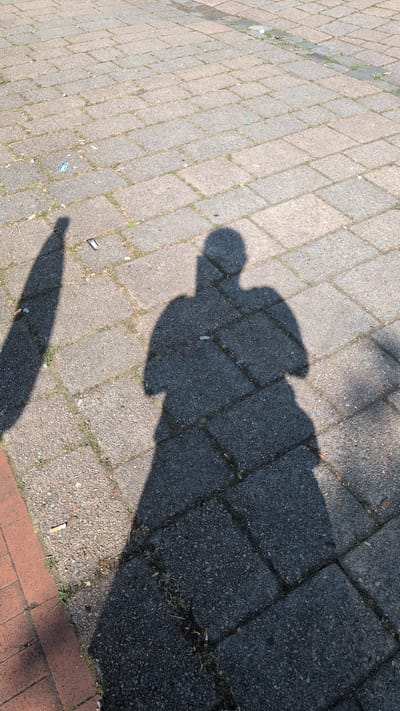 This is a photograph of two shadows cast on a brick-paved surface. The main shadow on the right appears to be that of a person standing and holding a phone up to their face, as indicated by the shape of the head and outstretched arm. A second, thinner shadow is visible to the left, suggesting another person or object. The ground is composed of rectangular paving stones, with some sparse vegetation growing in the cracks. Several cigarette butts are scattered across the paving stones, particularly in the foreground and left side of the image. The lighting suggests it is daytime, with the sun casting long shadows. The scene appears to be an outdoor public space, possibly a sidewalk or plaza, in the city of Osnabrück, Germany, given the characteristic style of paving stones often found in European cities.