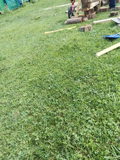 The image depicts a grassy yard with various construction materials and tools scattered across it. In the background, a green wooden fence is visible, suggesting a rural or suburban setting. A partially assembled wooden structure, consisting of stacked logs and planks, stands in the middle ground. A red wheelbarrow is positioned near this structure. Additionally, a blue shovel and a yellow tool are lying on the grass. The presence of these items indicates that some form of outdoor construction or DIY project is underway. The overall environment is a sunlit day with ample green grass. The specific location is identified as Islambaevo, Russia, though this information is not visually verifiable from the image itself.