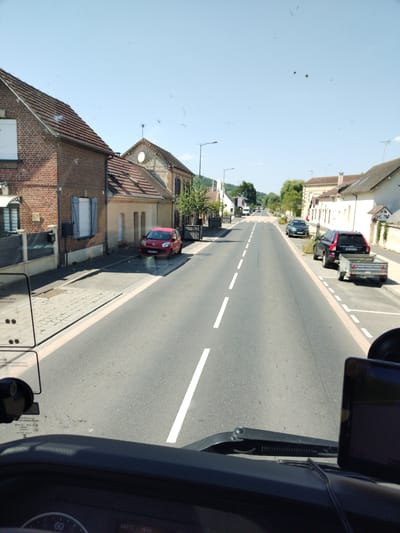 The image shows a street scene in Acquigny, France. The viewpoint is from inside a vehicle, looking forward. The road is asphalt, with dashed white lines marking the lanes. On the left side of the road are houses with brick and stone facades, red tile roofs, and some with blue shutters. A red car is parked on the side of the road. On the right side of the road are more houses, trees, and parked vehicles, including a black SUV towing a small trailer. The sky is clear and blue. There are some minor smudges on the windshield, creating a few indistinct dark spots in the sky and on the houses. The overall environment suggests a sunny day in a small town or village.