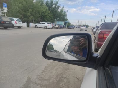 The image captures a street scene in Denisovka, Kazakhstan. The foreground shows the side mirror of a white car, reflecting the sky and a person holding a smartphone. In the mirror's reflection, a road with other vehicles and trees is visible. Several parked cars, including a white station wagon and a black sedan, are present on the left side of the road. Further down the street, more cars are parked in front of a small blue building. A blue and white parking sign is also visible on the left. The environment appears to be an outdoor, possibly semi-urban or suburban area, with a paved road and a cloudy sky.