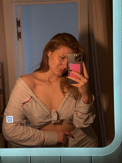 The image is a mirror selfie taken indoors, depicting a female subject from the chest up. The subject, a woman with light brown hair falling over her shoulders, is seen in her reflection. She wears a light-colored, possibly pale blue or white, pinstriped collared shirt with red piping accents, unbuttoned and tied at the waist, exposing her shoulders and midriff. Her right hand is placed on her lower garment or hip, while her left hand holds a smartphone, used to capture the image. The smartphone features a glittery clear case and a rectangular bright pink accessory with multiple circular indentations on its back. The subject's left hand displays dark nail polish. An illuminated white square icon with a power symbol is visible on the bottom-left reflective surface of the mirror. Below the mirror, a row of dark-colored tiles is visible. The reflected background includes a light-colored doorframe with two dark, possibly metallic, hinges on the left, and a dark curtain or fabric on the right. The overall lighting is warm and subdued. The image provides no discernable visual information to identify its location as Бакча-Сарай, Russia.