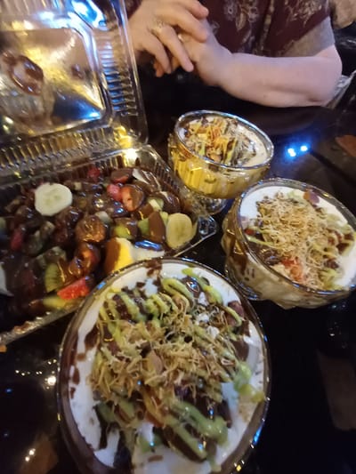 The image shows a close-up view of several desserts, possibly at a restaurant in Nematjan, Uzbekistan. In the foreground, a clear plastic container holds a medley of chopped fruits like strawberries, kiwis, and bananas, all generously drizzled with chocolate sauce. To the right and slightly behind, two glass bowls contain creamy desserts topped with what appears to be shredded coconut or a similar garnish, and drizzled with green sauce and chocolate sauce. A person's hands are visible in the upper right corner, with jewelry on one of the fingers, suggesting they are about to partake in the desserts. The table surface is dark and reflective, with subtle blue and yellow light reflections, hinting at ambient lighting. The overall scene conveys a sense of indulgence and enjoyment of sweet treats.