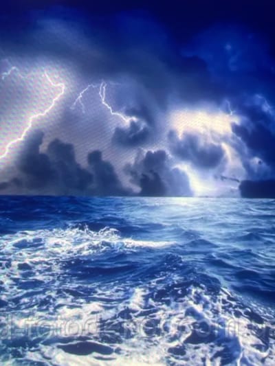 The image depicts a turbulent marine environment under a dark, stormy sky, with the scene purportedly located in the city of Zelenyi Hai, Ukraine. The upper half of the image features heavy, dark blue and gray storm clouds, actively illuminated by multiple prominent streaks of white and light purple lightning. These lightning strikes signify intense atmospheric electrical discharge. Patches of brighter light are visible breaking through or reflecting off the clouds in various sections, suggesting distant or obscured light sources behind the storm. The lower half of the image portrays a rough, dark blue sea characterized by numerous whitecaps and foamy waves in motion, filling the foreground and extending towards a distinct horizon line. The water shows dynamic movement, indicative of strong winds and rough conditions. No land features, vessels, people, or other specific objects are visible in the environmental scene. Faintly visible in the bottom section of the image is a watermark or text, "detorodka.com.ua".