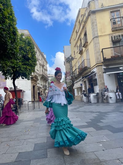 The image depicts a street scene in Jerez de la Frontera, Spain. A woman in a traditional flamenco dress with a large turquoise ruffled skirt and embroidered white top is the central figure. She wears a red flower in her hair. Behind her, there is a building with classical architectural details. Further back, people sit at tables outside a bar or restaurant. Another woman is partially visible in a similar flamenco dress with a purple skirt. A tree is present in the foreground. The ground is paved with grey stone tiles.