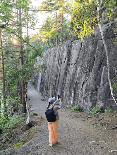 In Skriketorp, Sweden, a person wearing an olive-green jacket, light blue baseball cap, and peach-colored pants walks along a gravel path beside a large, dark gray rock face. The person is holding a smartphone in their right hand, taking a photograph of the rock face. A navy blue backpack is worn by the person. The path is bordered by trees on the left, with dappled sunlight filtering through the leaves. The rock face is textured with various cracks and lighter-colored mineral deposits. In the distance, the path curves out of view, and other people can be seen walking. The environment appears to be a natural park or trail.