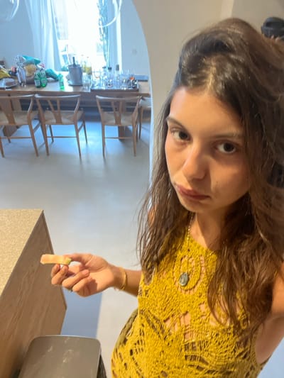 An indoor photograph depicts a young female in what appears to be a residential or hospitality setting in Agios Ioannis Diakoftis, Greece. The subject, a female with long, dark hair, wearing a yellow crocheted or knitted sleeveless top and a gold-colored necklace with a dark pendant, occupies the right foreground. She holds a light-colored, elongated food item, possibly a piece of fruit, in her right hand, extended over a light-colored, speckled countertop. Her facial expression exhibits slightly downturned lips and a direct gaze towards the camera.
In the background, a spacious, brightly lit room is visible, featuring a long, light-toned wooden dining table surrounded by multiple light-colored wooden chairs with woven seats. The table is set with a silver ice bucket, an assortment of clear drinking glasses, several green bottles, and a large clear bowl containing snacks. Natural light permeates the space from a large window adorned with sheer curtains. The architectural elements include light-colored walls and flooring, and an arched opening leading to an adjacent area. Transparent, spherical decorative objects are suspended from the ceiling near the window. A dark, unidentified object is partially visible below the countertop in the immediate foreground.