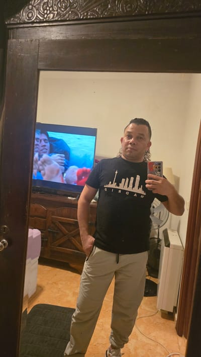 A man is taking a selfie in a mirror inside a room. The man is wearing a black t-shirt with a graphic design and light-colored pants. His phone has a colorful case. Reflected in the mirror is a television displaying a scene involving a person. A fan and a white device are located behind the man. The floor is tiled in what appears to be a reddish-brown color. The exact location within Real Sitio de San Ildefonso, Spain is not discernible from the provided image.