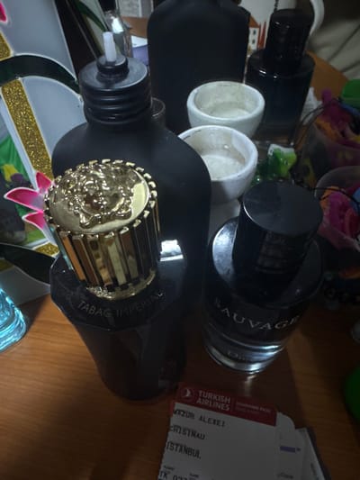 In Ciorescu, Moldova, the image displays a collection of personal items situated on a light brown wooden surface. Three primary perfume bottles occupy the midground. Dominating the left is a dark, possibly black, bottle labeled "TABAG IMPERIAL" with a prominent ornate golden cylindrical cap featuring a detailed embossed design. To its right is a clear glass bottle containing a dark liquid, identified as "SAUVAGE" by "DIOR", capped with a black lid. Partially visible behind and to the left of the "Tabag Imperial" bottle is another black matte bottle, equipped with a black threaded cap and a white plastic nozzle. Behind these bottles, two small, light-colored, possibly ceramic, bowls are arranged. A larger, dark-colored bottle is partially visible further back. In the lower right quadrant of the image, a Turkish Airlines boarding pass is present, detailing the passenger as "MAZUR ALEXEI" with destinations "CHISINAU" to "ISTANBUL" and flight number "TK 037". Additional objects in the background include a blue-tinted glass item, a decorative element with white and gold glittery segments and a pink floral detail, and various indistinct colored fabrics or objects suggesting a personal environment.