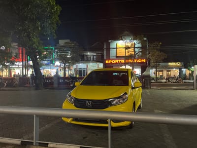 The image depicts a street scene at night in Potroyudan, Indonesia, with a bright yellow car parked in the foreground. The car, a Honda Brio, is the central focus, positioned on a paved surface. Behind the car, buildings are visible, illuminated by neon signs and lights. One sign prominently displays "SPORTS STATION" in glowing orange letters. Other businesses, such as a place called "Ma'Rici," are also discernible. Trees with foliage are scattered throughout the scene, and power lines crisscross overhead. Several people are present, some standing and others walking, and a few motorcycles are parked in the background, suggesting a lively commercial area. The overall impression is of a well-lit urban environment at night.