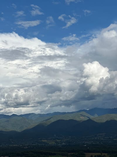 An elevated landscape photograph captures a mountainous vista within the city of Ban Mai Saha Sampan, Thailand. The upper two-thirds of the frame consists of a partially clear blue sky featuring extensive white and grey cumulus and stratocumulus cloud formations. A singular, small, dark, unidentified aerial object is visible in the upper mid-right blue sky. The lower third depicts a layered mountain range densely covered in green and dark green foliage, with pronounced areas of sunlight and deep shadow. On a sunlit mid-ground mountain slope, a cluster of structures with reddish roofs is present. Below the immediate mountain slopes, a valley floor extends into the distance, showing a mosaic of dark forested areas, lighter green agricultural fields, and scattered small settlements. No human figures are visible.