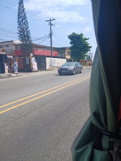 A street scene in Animashawun, Nigeria, is captured from inside a moving vehicle, with a portion of the vehicle's interior and a green tarp obscuring the right side of the frame. The asphalt road features double yellow lines indicating the lane dividers. A grey sedan, license plate KAV 596A, is traveling in the oncoming lane, its headlights on. Several pedestrians are visible on the sidewalk in the distance. Buildings line the street, including one with a red facade and a tall, slender evergreen tree in front of it. Another building has a lush green tree with a wider canopy. Utility poles and a dense network of overhead wires dominate the sky. Signs and posters are displayed on some of the buildings and on freestanding stands along the sidewalk. The sky is partly cloudy.