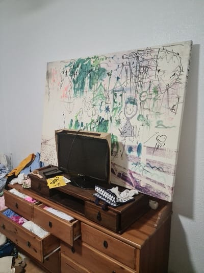The image displays an indoor scene in a city in El Campello, Spain. A large, white canvas painting, covered with abstract, childlike drawings and scribbles in green, pink, black, blue, and purple, including stick figures and a smiley face, leans against a plain white wall.

In front of the painting, a light-brown wooden dresser with multiple drawers is positioned. Three of the upper drawers are partially open, revealing various folded clothes and fabric items within. On top of this dresser, a darker brown wooden TV stand holds a small, black, flat-screen television, which is powered off. A black cable extends from behind the television.

To the left of the television, a small, dark-brown wooden box with a decorative metal clasp is visible, holding a yellow card with the word "GOLF" printed in black. To the right of the television, a rectangular container with a blue and white patterned fabric cover, likely a tissue box, is present next to a small, white cylindrical object.

The area to the left of the dresser is cluttered with a pile of clothes, including yellow and orange garments. Additional items are partially visible on the floor in the lower left corner. No individuals are present in the scene, and no actions or interactions are depicted.