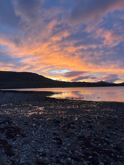 The image depicts a coastal scene at dusk, with a dramatic sky filled with clouds illuminated by the setting sun. The sky transitions from deep blues and purples at the top to vibrant oranges and pinks closer to the horizon. A silhouetted mountain range with rolling hills forms the backdrop, with a few scattered houses visible along the coastline. The foreground is a rocky beach, with dark, wet stones and patches of seaweed. The water in the middle ground is calm and reflects the colors of the sky. The overall impression is one of natural beauty and tranquility. The location is identified as Sconser, United Kingdom. There are no people or animals visible in the image.