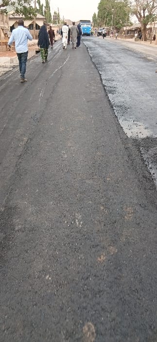 A newly paved asphalt road is seen at an angle. The asphalt on the upper side of the road appears fresher and still wet, while the lower side is dry and already set. Several individuals stand near a truck in the distance, partially obstructing the road. One person is visible in the foreground on the left. Buildings and trees line the sides of the road.