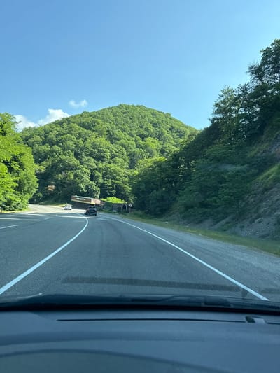 The image depicts a view from inside a vehicle looking out at a winding asphalt road through a lush, green, mountainous landscape under a clear blue sky. The road has white lane markings. On the left side of the road, there is a dense forest covering a hillside. On the right side of the road, a steep, rocky embankment is covered with vegetation. In the distance, a building with signs in Cyrillic script appears to be a roadside establishment. Two vehicles are visible on the road ahead: a white car and a black SUV. The scene is serene and natural, suggesting a rural or mountainous environment. It is not possible to determine the exact location of Tyumenskiy, Russia from the provided image.