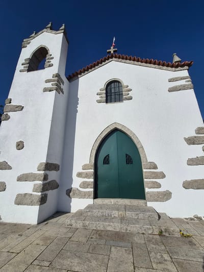 The image shows the side view of the Igreja da Misericórdia de Esposende, located in Esposende, Portugal. The church has a white exterior with gray stone accents. The front door is green and arched. There is a small, barred window to the left of the door. The roof is red tile. A bell tower is visible on the left side of the church. The sky is blue. The image is taken from a low angle and rotated counterclockwise. There are stone steps visible to the right of the door.