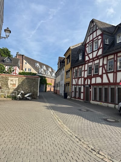 The image depicts a narrow cobblestone street in Wetzlar, Germany. The street is lined with traditional German timber-framed buildings, characterized by their white infill panels and dark wooden beams. Some buildings have multiple stories with numerous windows. To the right of the street, there are manhole covers and a shadow cast by an unseen object. In the distance, more buildings can be seen, including one with a red brick facade and multiple dormer windows. On the left side of the street, near the middle of the frame, two white scooters are parked next to a stone wall. The sky is blue with wispy clouds, suggesting it is daytime. The scene is quiet and appears to be a typical European street on a clear day. There is a street lamp visible in the lower left corner of the image. No people are visible in the image.