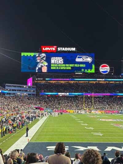 The image shows a view of Levi's Stadium during a football game. A large scoreboard displays an advertisement for Jason Myers, highlighting his record for most field goals made in a Super Bowl, next to the Seattle Seahawks logo. A Pepsi advertisement is positioned above. The football field is visible with yard lines and goalposts, and the stands are filled with spectators. The scene is captured at night.