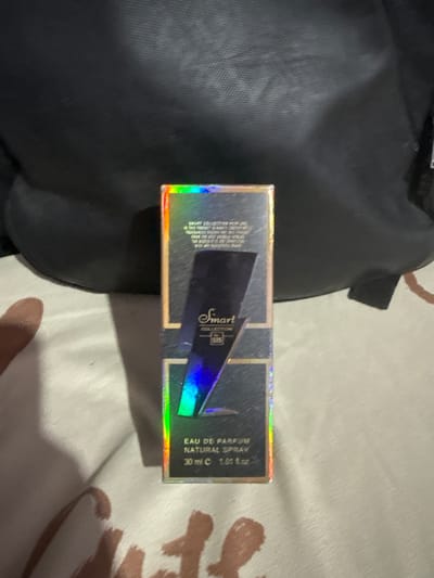 A close-up shot shows a small, rectangular perfume box with holographic accents. The box is positioned on a textured surface, possibly fabric, with a dark, somewhat blurry object in the background. The perfume box itself is predominantly dark blue with gold lettering and iridescent highlights. Text on the box includes "Smart Collection", "EAU DE PARFUM", "NATURAL SPRAY", and "30 ml e 1.01 fl.oz". The number "335" is also visible. The image does not provide any information about its location in Manila, Philippines, or any details about people, actions, or interactions. The scene is static and focuses solely on the product.