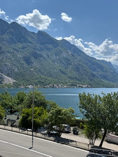 A landscape view taken at Donji Orahovac, Montenegro shows a bright, clear sky with scattered white clouds against a blue backdrop. On the left is a steep, mountainous terrain covered with dense green trees. A small town rests at the base of the mountain, running parallel to the body of water, Kotor Bay. On the right, the shot includes the edge of a paved road with white dividing lines and some parked vehicles. Trees are positioned between the road and the water. A tall lamppost stands in the middle of the frame near the roadside, adding to the scenic depiction of the location.