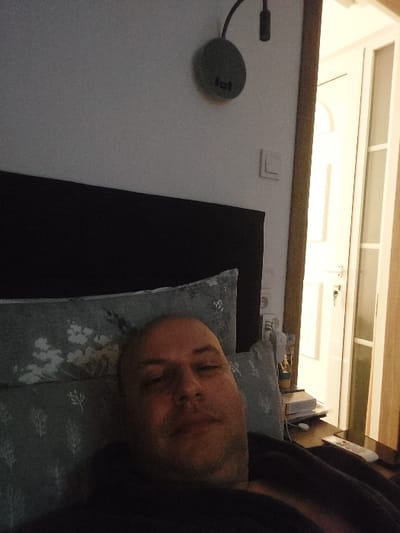 The image depicts an adult male in a bedroom located in Kalyvia Thorikou Municipal Unit, Greece. The man, bald with visible stubble, lies in bed, appearing to take a selfie. He is supported by two light blue-grey pillows patterned with white botanical designs, and is partially covered by a dark fabric. A dark headboard frames the bed. To the left, a white wall features a circular dark grey wall-mounted lamp with an articulated arm and a white rectangular light switch below it. To the right of the bed, a dark wooden nightstand holds stacked items and a white remote control. Further right, a white door, possibly with glass panels, is partially open, revealing a lighter area beyond and a key in its lock. The scene is dimly lit.