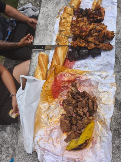 The image shows an outdoor picnic scene. In the foreground, a white paper wrapper is spread on a concrete surface. On the wrapper, there is a pile of chopped meat, a piece of cooked plantain, and some red sauce. To the left, a white plastic bag contains more bread. In the background, a person's arms and legs are visible, sitting on a stone ledge. Another person's legs and hand, holding a piece of bread, are also visible on the left side of the image. A knife is resting on the paper wrapper. Further back, a stack of grilled chicken skewers and sliced bread are laid out on paper. The overall setting appears to be a casual outdoor gathering. The text "Campezo / Kanpezu, Spain" is not present in the image or its crops, and therefore cannot be included in the description.