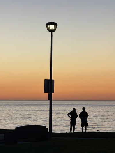 A silhouette of a lamp post with an illuminated head and a square sign is centrally placed. The background features a gradient sky transitioning from light gray to orange, bisected by the line of water. Two silhouetted figures stand near the water's edge, one with hands on hips. A bench and patch of grass are partially visible on the right.