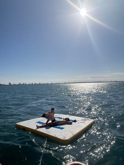 The image depicts an open-water scene under a bright sun and clear blue sky, offshore from Veracruz City, Mexico. Two shirtless males are situated on a rectangular white and yellow inflatable water platform floating on choppy blue-green water. Sun glare creates a bright reflection pattern on the water's surface to the right. In the distant background, a low-rise city skyline is visible along the horizon.
One male lies supine on the platform, wearing dark patterned swim shorts. His head rests on a blue towel or mat, and his arms are positioned behind his head. A second male sits upright behind the first, also wearing dark swim shorts, looking towards the viewer with a slight smile. A dark line or rope extends from the front of the inflatable platform, leading out of frame towards the lower right. A small white object, possibly a bird, is visible in the upper left sky.