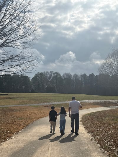 Three individuals are walking on a paved path alongside a grassy area, with trees in the background. The person in front is wearing a white shirt, the middle person is wearing jeans, and the last is wearing a black shirt. Their shadows stretch ahead of them on the path. The sky is filled with clouds.