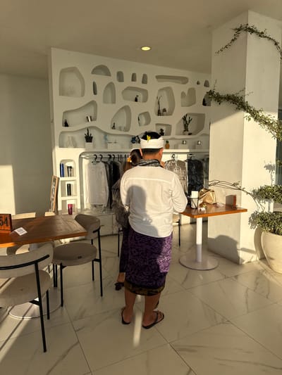 The image, taken in Kedisan, Indonesia, shows a man inside a retail space. He is wearing a white shirt, a purple sarong, and a traditional white headdress with a flower. He is wearing flip-flops. He appears to be examining items on a clothing rack inside the shop. The shop's interior is characterized by a white wall with built-in display niches, some containing decorative items. A wooden table with chairs stands in the foreground. Above, there is foliage decorating the space. The floor is light-colored tile.