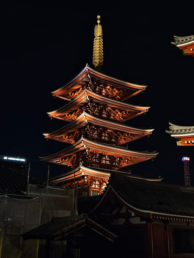 The image shows a nighttime view of the Senso-ji temple in Taito, Japan. The frame is dominated by the upward angle view of the five-story pagoda, its dark red and white detailed roofs contrasting against the black sky. The pagoda's finial is visible at the left. The architecture of an adjacent temple building is visible to the right, also lit up. Above and to the left of the pagoda, sections of a banner can be seen. In the upper center of the image, an observation tower with colorful lighting is just visible.