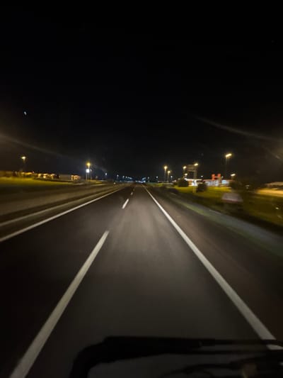 The image depicts a nighttime scene from the perspective of a vehicle traveling on a multi-lane highway in the city of Cerignola, Italy. The dark asphalt road surface is illuminated by the vehicle's headlights and numerous streetlights lining both sides of the roadway. White dashed and solid lane markings are visible, guiding traffic. On the immediate right side, a grassy verge transitions into an area with multiple structures, illuminated by bright artificial lights, consistent with a service station or rest area. These structures include what appears to be a canopy over fuel pumps and low-rise buildings. On the left side, beyond a similar grassy verge, a cluster of distant lights suggests further commercial or industrial development. The sky above is dark, indicating night conditions. No individuals or other vehicles are clearly discernible on the road. Slight motion blur is present, consistent with a moving vehicle. The bottom edge of a vehicle's dashboard or windshield frame is visible in the foreground.