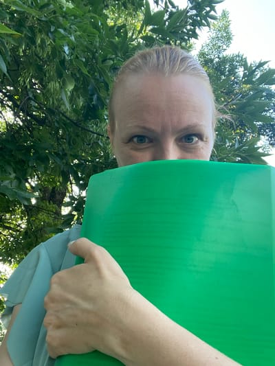 An adult Caucasian female with blonde hair pulled back is partially obscured by a bright green folder held in front of her face, with only her eyes and forehead visible. She is wearing a light blue, possibly silk, top. The woman is outdoors, with lush green foliage and branches of a tree forming the background. The sunlight filters through the leaves, creating a dappled effect. The image appears to have been taken at a slightly upward angle. There are no other people or objects in the immediate vicinity. The location is not identifiable as a specific part of Zapadny, Russia, but the environment suggests a park or garden.