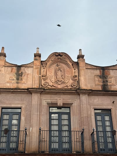 The image shows a view of the façade of a building in Zacatecas City, Mexico, under a cloudy sky. A single bird is flying in the upper center of the image. The building features pinkish-orange stone construction with ornate architectural details. Centered on the upper part of the façade is a bas-relief carving depicting the Virgin Mary, with the inscription "AVE MARIA" below it. Flanking this carving are decorative pilasters and finials. Below the ornate upper section are three windows, each with dark blue double doors and glass panes covered by sheer white curtains. Each window has a wrought-iron balcony with decorative scrollwork. A small potted plant is visible on the balcony to the left. The building exhibits signs of age and weathering, with some darker streaks on the stone.