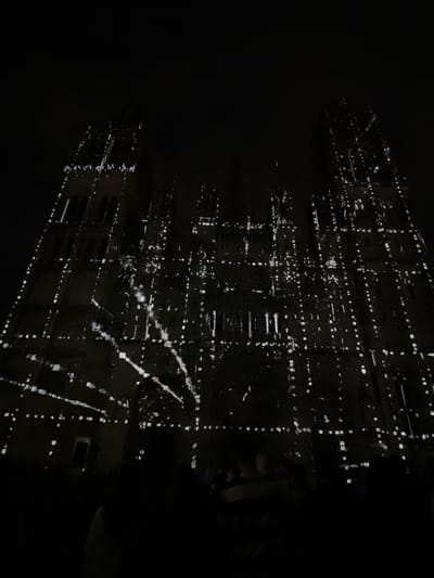 The image depicts a large Gothic cathedral, identifiable as Rouen Cathedral in Rouen, France, at night. The cathedral's facade is illuminated by complex white light projections creating a dynamic, grid-like pattern of lines, dots, and abstract shapes that appear to animate across its stone surface. Multiple architectural towers and spires are visible against the dark, cloudless sky, their original details mostly obscured by the darkness and the projected light. In the foreground, at the bottom of the frame, several dark, indistinct human silhouettes are visible, presumably spectators observing the light display. The overall scene is dark, with the illuminated cathedral being the sole significant light source.