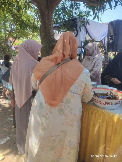 Three women wearing hijabs stand by a table at an outdoor event. The woman closest to the camera wears a floral dress with a light orange hijab and a shoulder bag. Another woman beside her wears a light purple hijab. A third woman with a matching hijab is visible behind them near a wedding tent. A large bowl with food sits on the table in front of the woman in the floral dress. A speaker and other individuals are visible in the background.