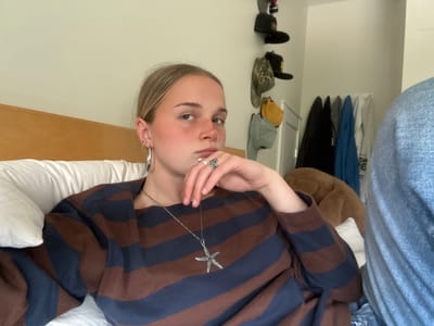 A young person with blonde hair is lying on a bed with a white pillow and a wooden headboard. The person is wearing a brown and blue striped shirt and has a silver starfish brooch attached to the chest of the shirt with a silver chain extending to the person's hand, which has a silver ring on it. The person has multiple piercings in the left ear and a nose piercing on the left side. In the background, there is a closet with clothes hanging inside and a rack of hats on the wall. It is not possible to discern the location within the city of Squamish, Canada, based on this image.