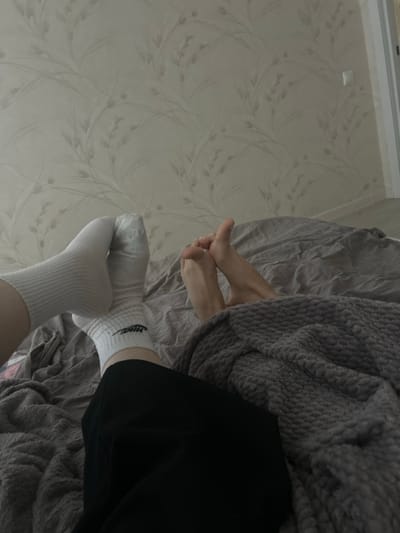 The image depicts a close-up view of two pairs of feet on a bed. One pair of feet is wearing white athletic socks, one of which has a Nike logo. The other pair of feet is bare and appears to be intertwined with the first pair. Both pairs of feet are positioned on a gray textured blanket. In the background, a bed with gray linens is visible, and beyond that, a wall with a floral wallpaper pattern. A light switch is mounted on the wall to the right. The scene is indoors, suggesting a domestic setting. The image does not provide specific information about the location within Veshki, Russia, beyond it being an interior residential space.